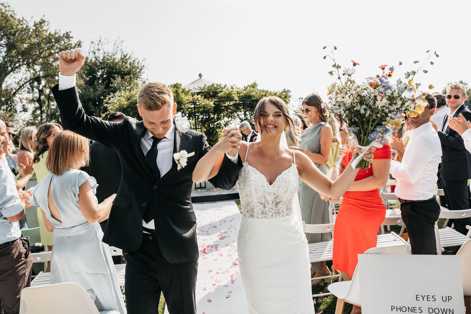 Excited newlyweds celebrate walking down the aisle with raised arms at outdoor garden wedding.