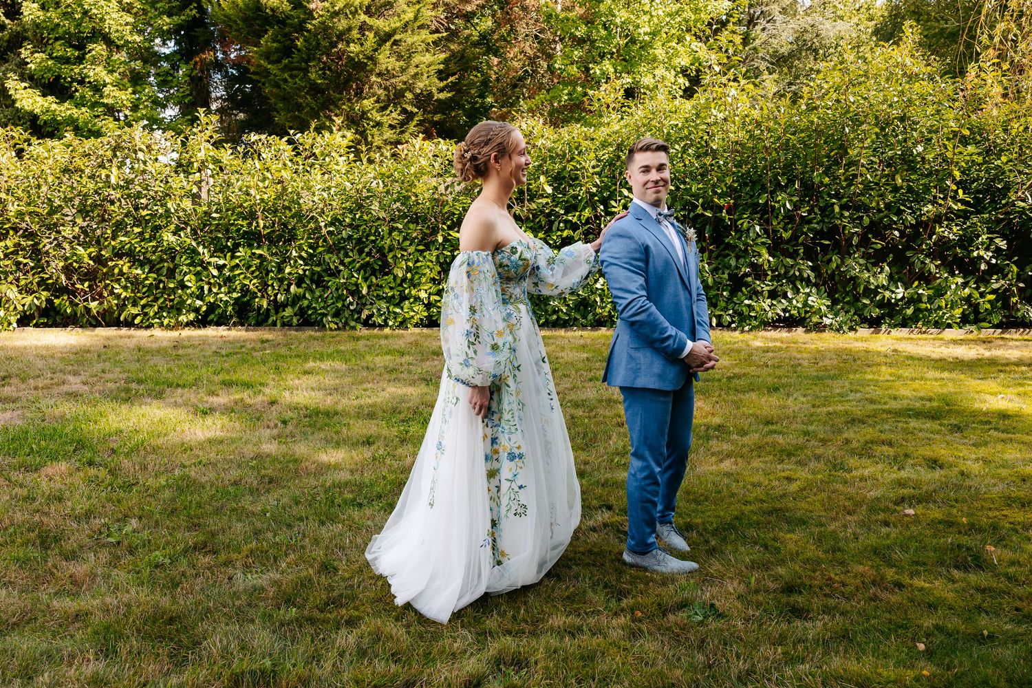 Bride in floral wedding dress and groom in blue suit hold hands while walking on garden lawn.