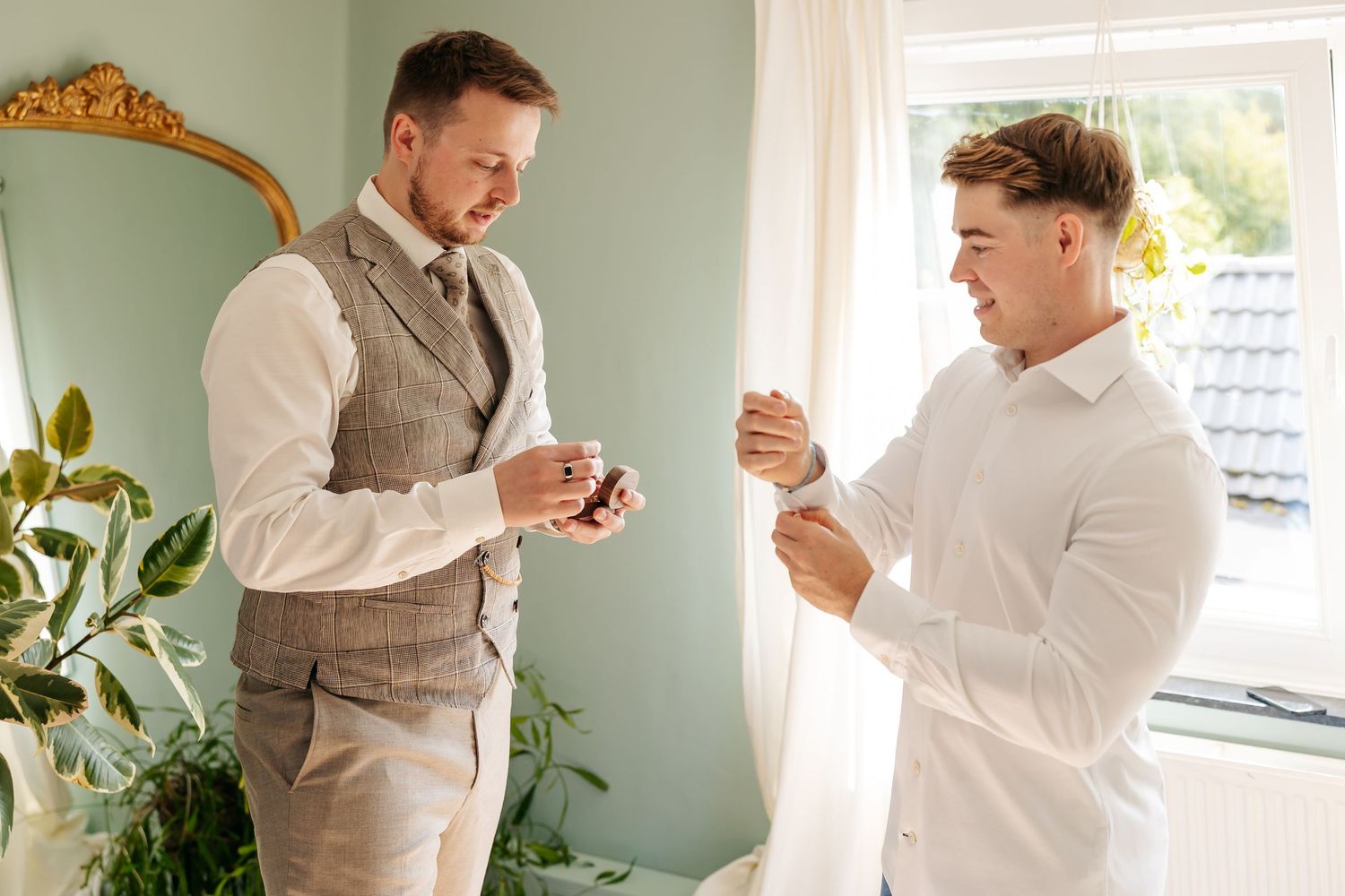 Two groomsmen in suits examining wedding rings and cufflinks while getting ready indoors.