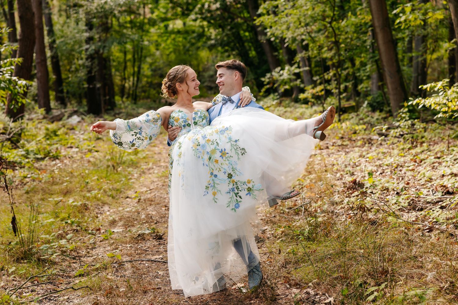Groom lifting bride in a floral wedding gown while spinning in a sunlit forest setting.