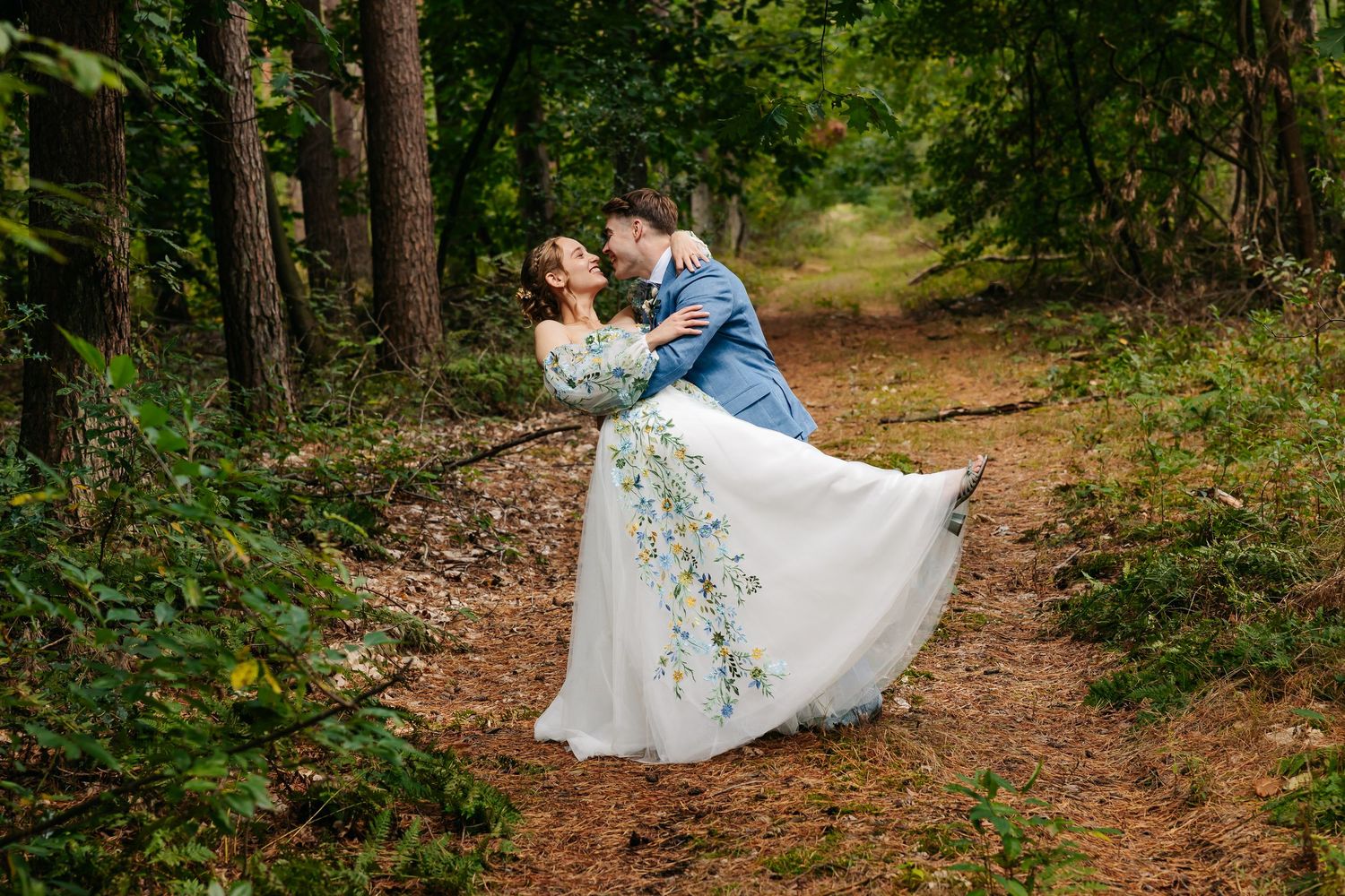 Groom dipping bride in floral wedding dress on forest trail.