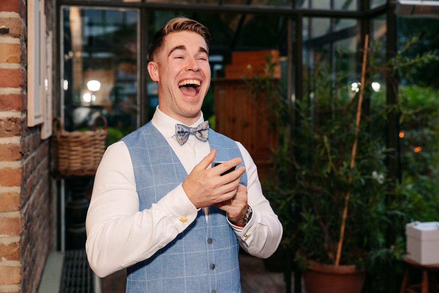 Laughing groom in blue checkered vest and bow tie at wedding reception.