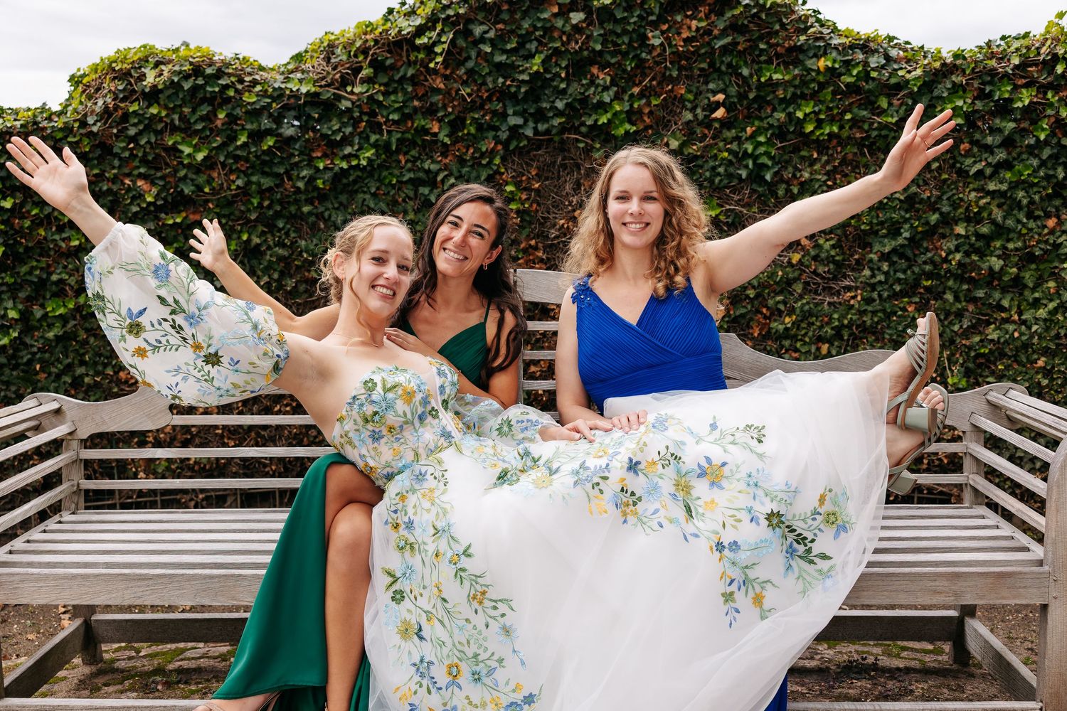 Bride in floral gown celebrating with two bridesmaids in green and blue dresses on garden bench.