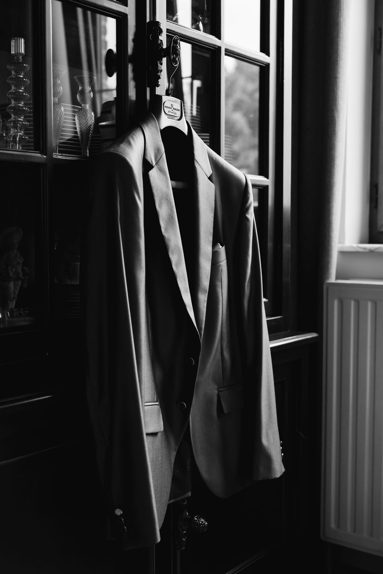 Black and white photo of grooms suit jacket hanging on wooden hanger near window.