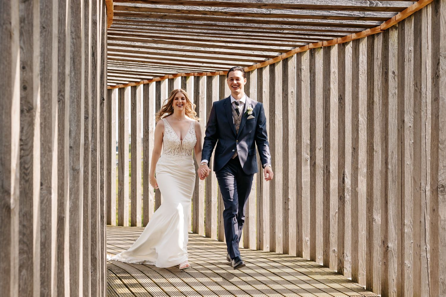 Bride and groom smiling and walking hand in hand through a sunlit rustic wooden pergola.