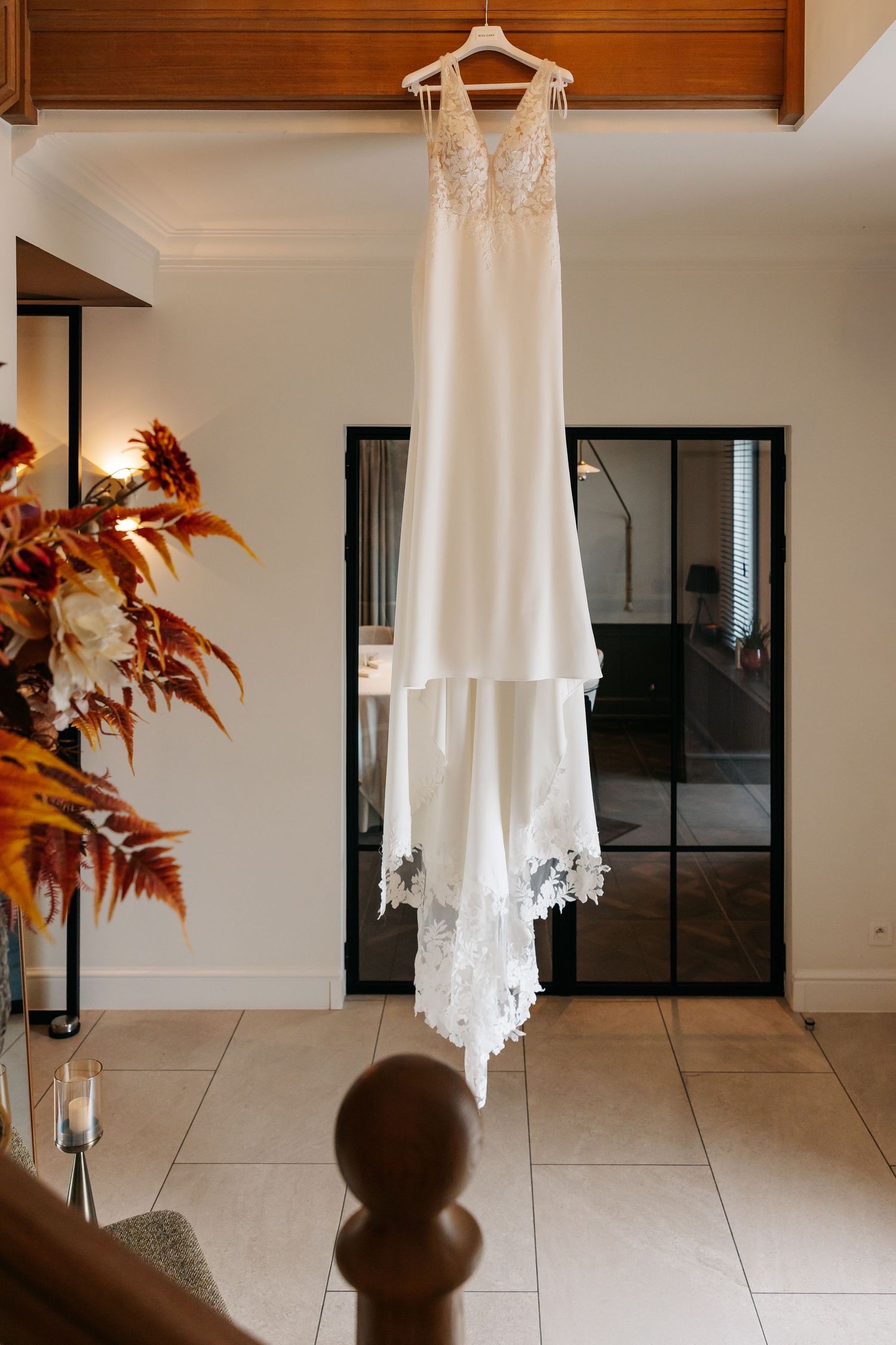 Elegant white wedding dress with lace hem hanging on a hanger in a bright modern room with autumn floral decor.