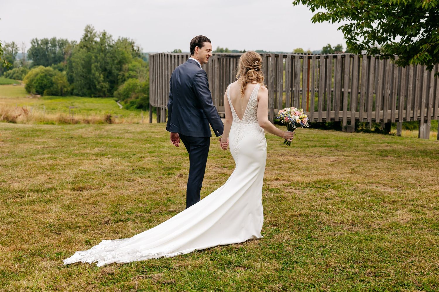 Bride in elegant backless white gown with long train walking with groom in green countryside setting.