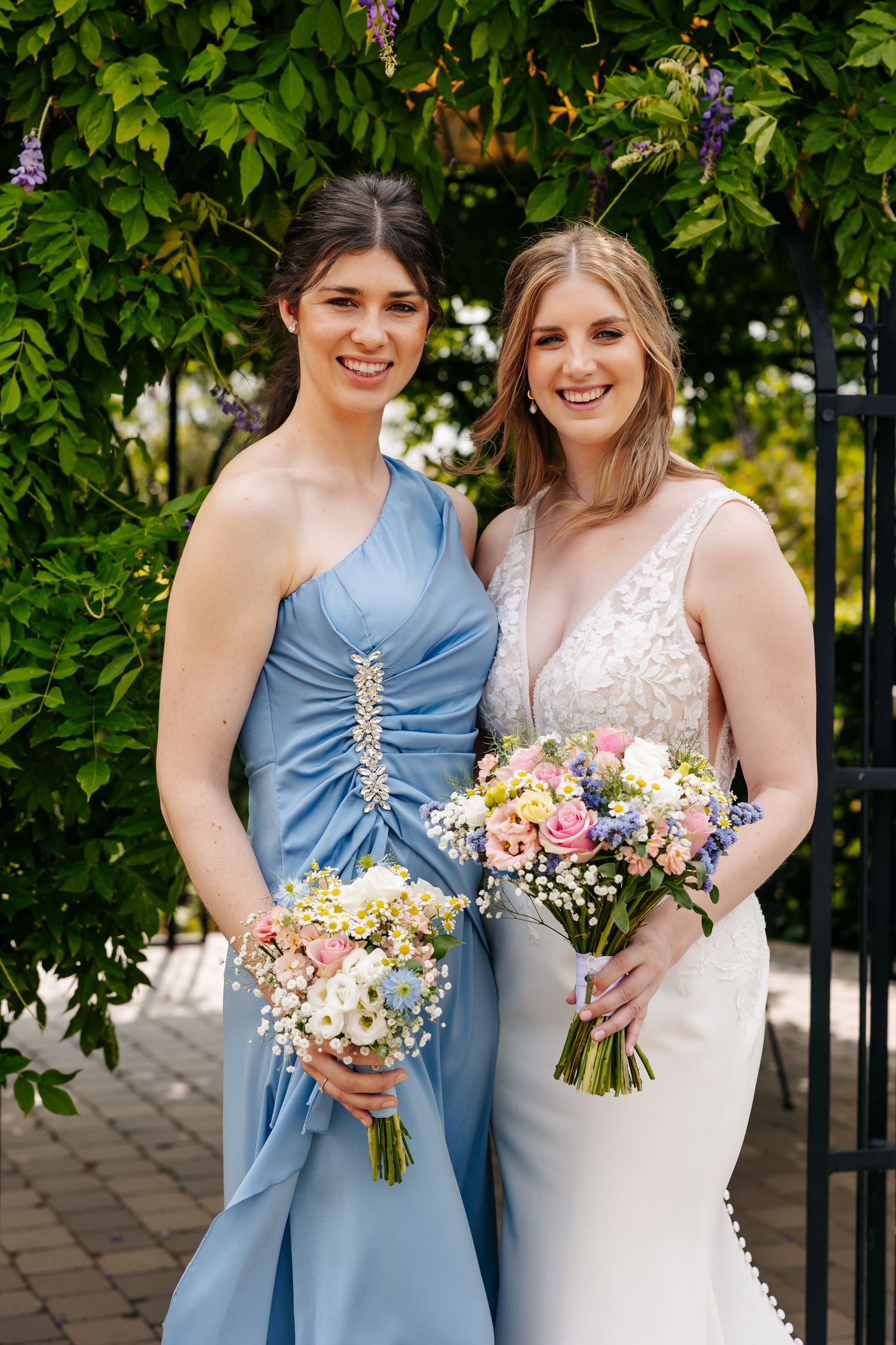 Bridesmaid in blue one-shoulder dress and bride in white lace gown holding bouquets by garden gate.
