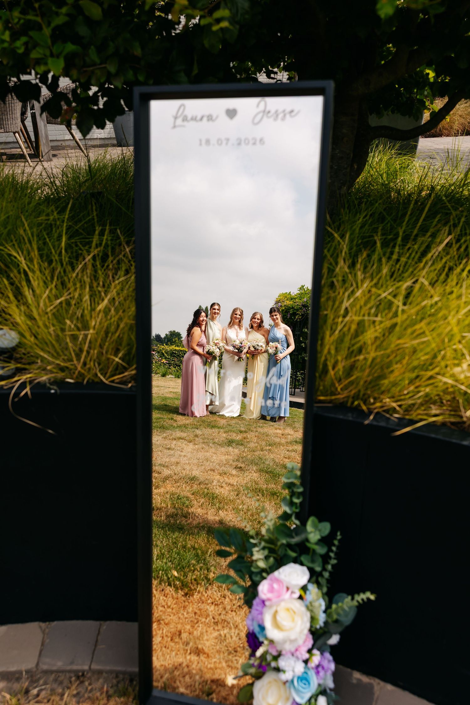 Wedding mirror sign for Laura and Jesse reflects bride and bridesmaids posing on a sunny lawn outdoors.