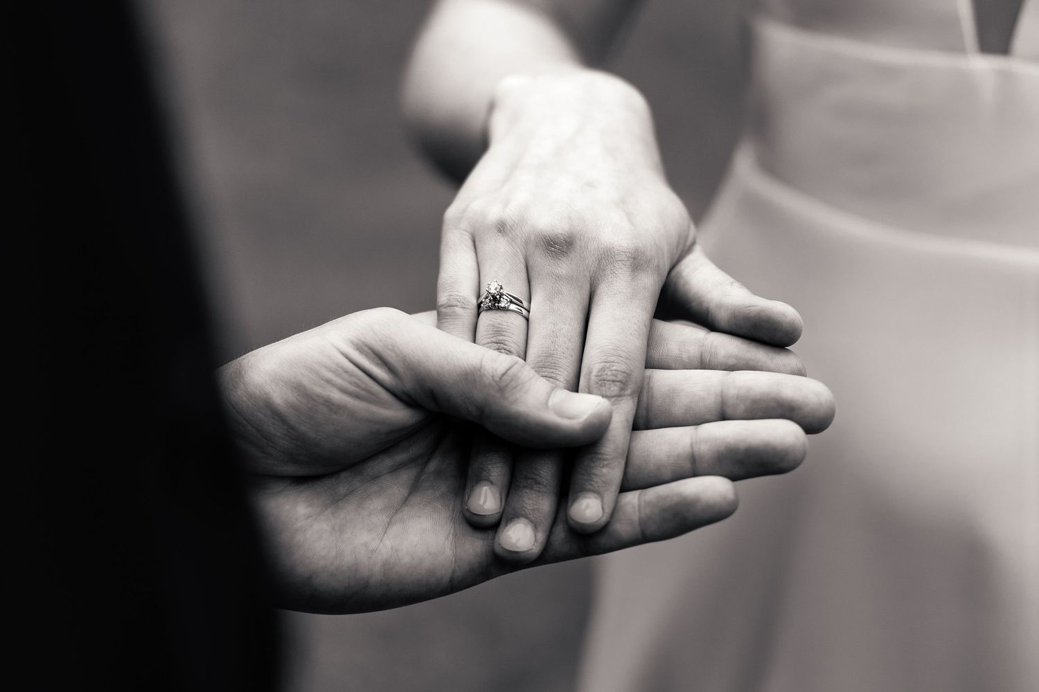 Close-up black and white photo of bride and groom holding hands with engagement ring visible.