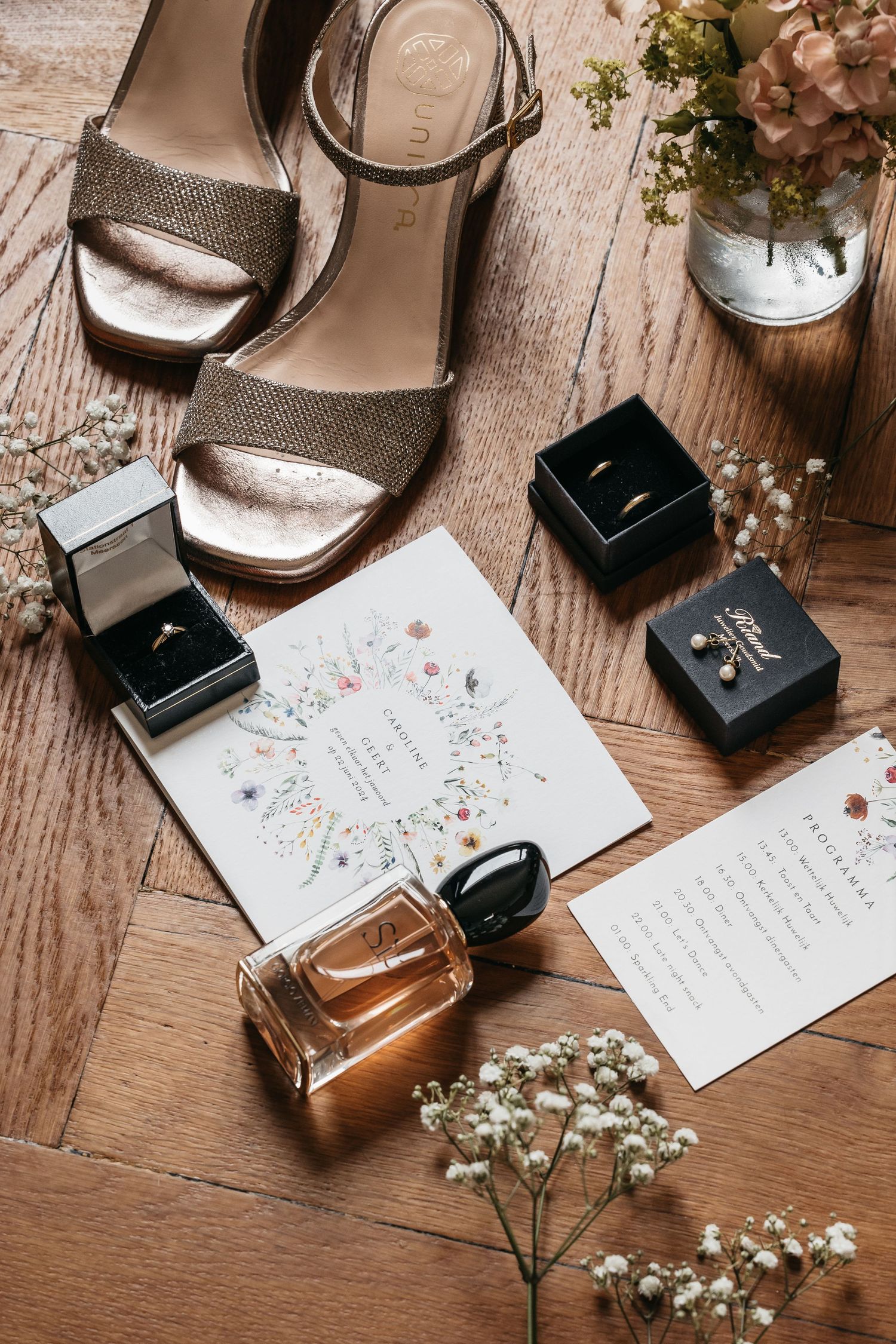 Wedding flat lay with gold heels, rings, perfume, and floral invitation on wood floor.