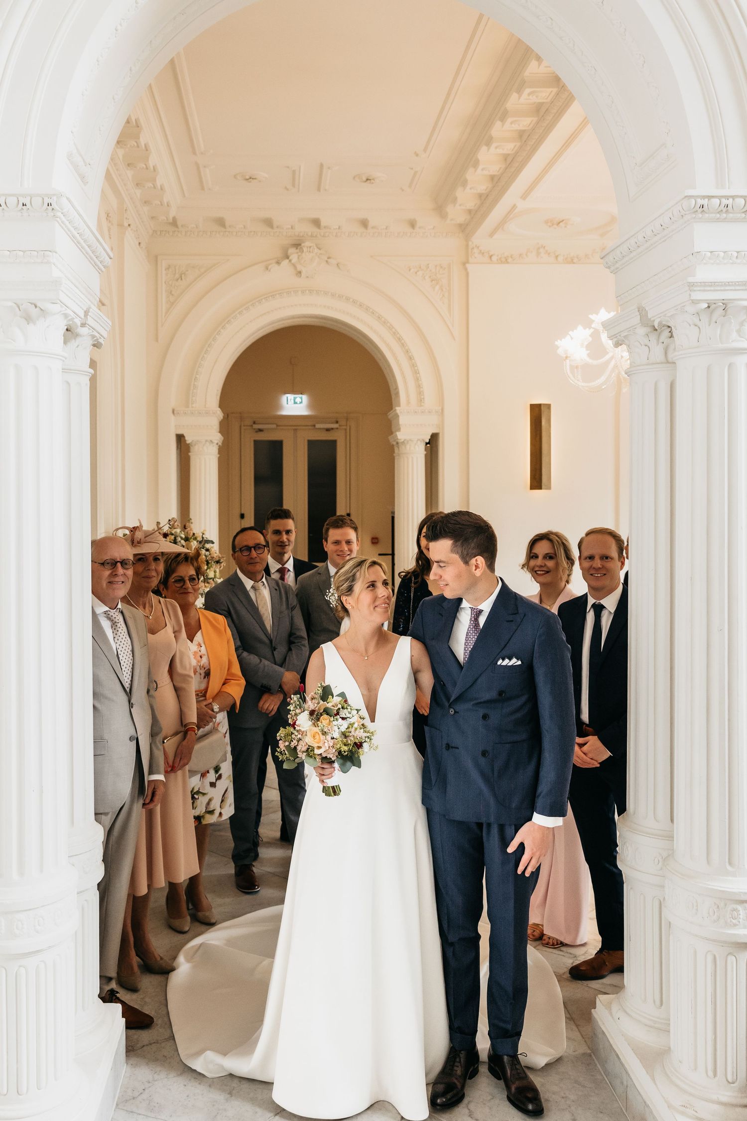 Bride and groom walk through elegant white arched hallway surrounded by wedding guests after ceremony.