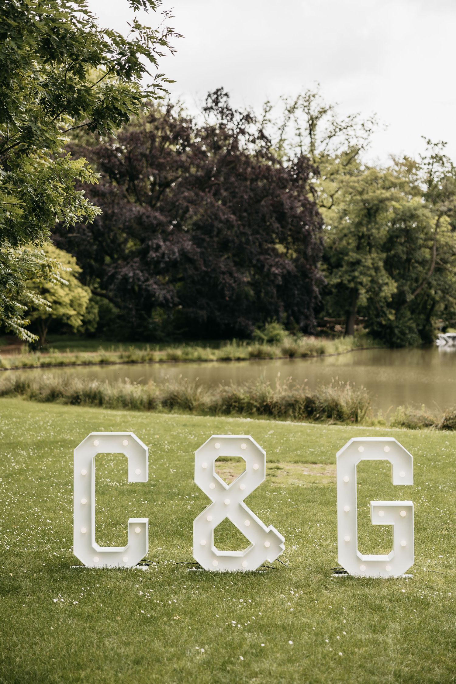 Illuminated marquee letter signs spelling C&G on green lawn with trees and pond in background at wedding venue.