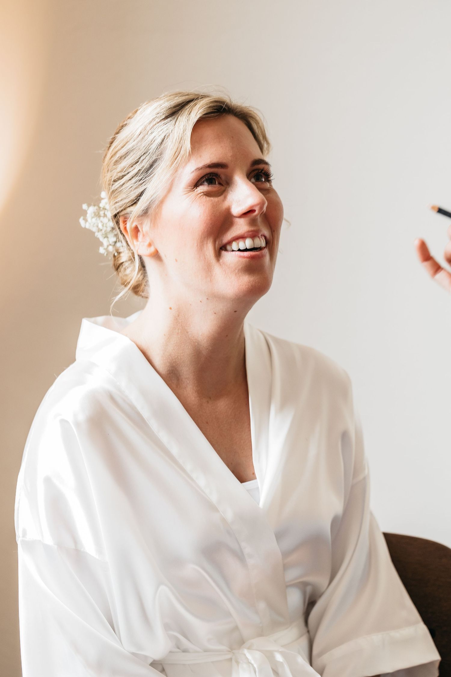 Radiant bride in white silk robe with floral hair accessory laughs joyfully during wedding preparations.
