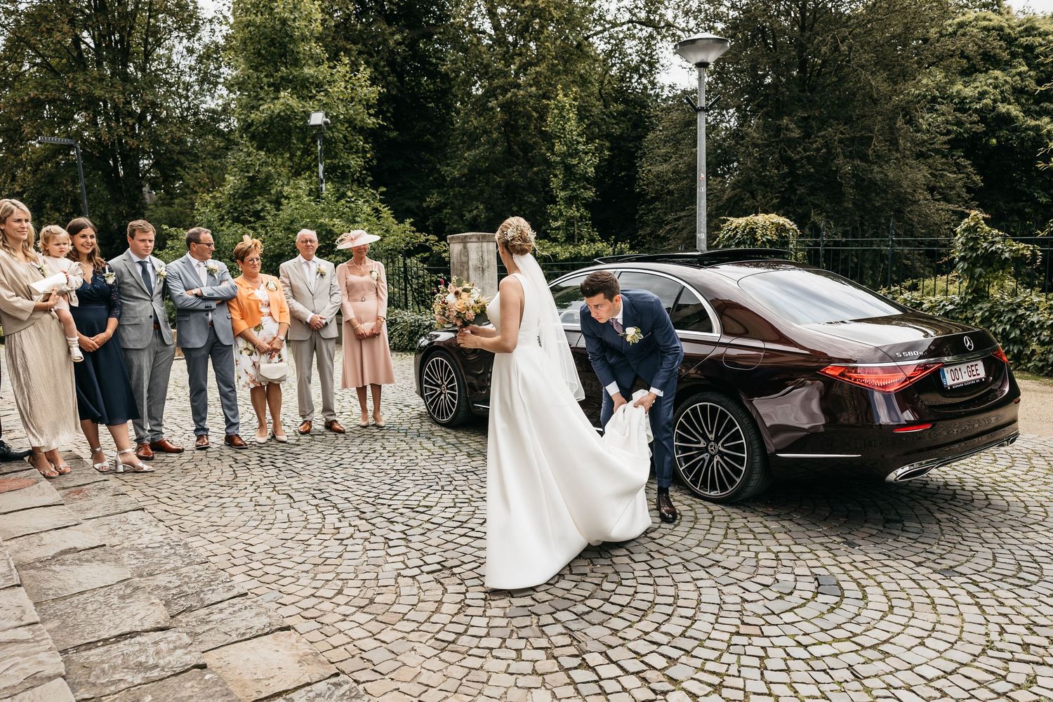 Groom adjusting brides white wedding dress train beside a black Mercedes while guests watch outdoors.