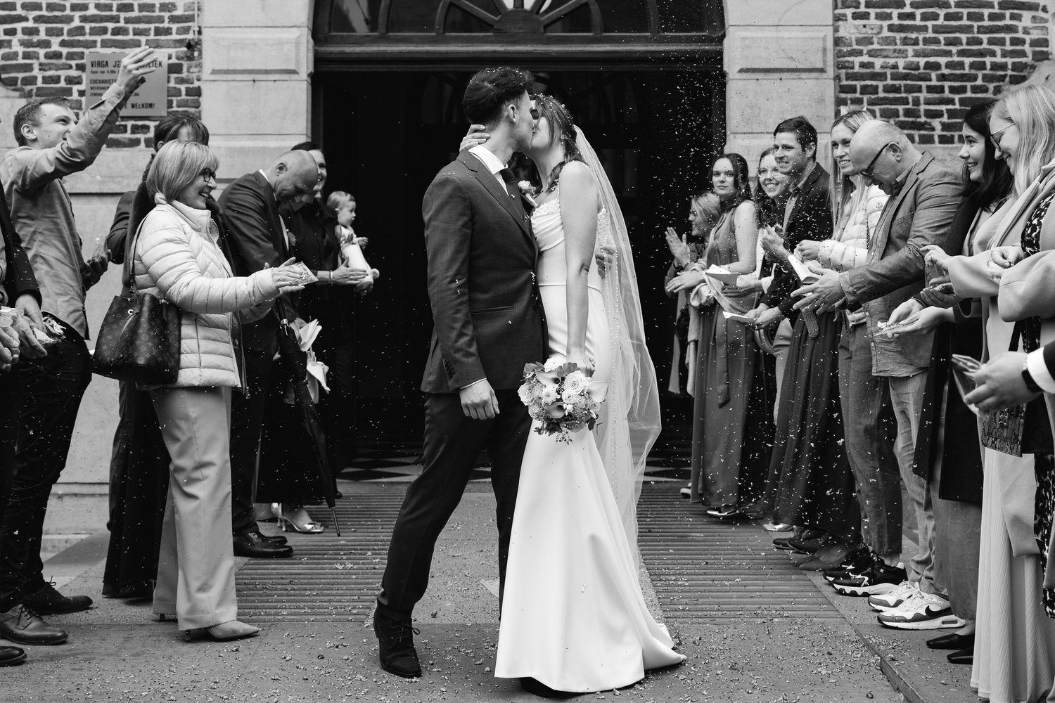 Newlyweds share a kiss outside church as guests toss confetti in black and white photo.