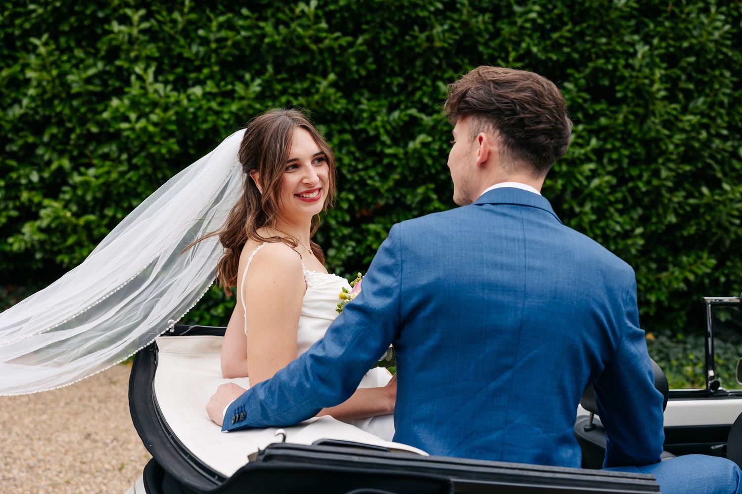 Smiling bride wearing veil looks at groom in blue suit while seated in classic white convertible car.