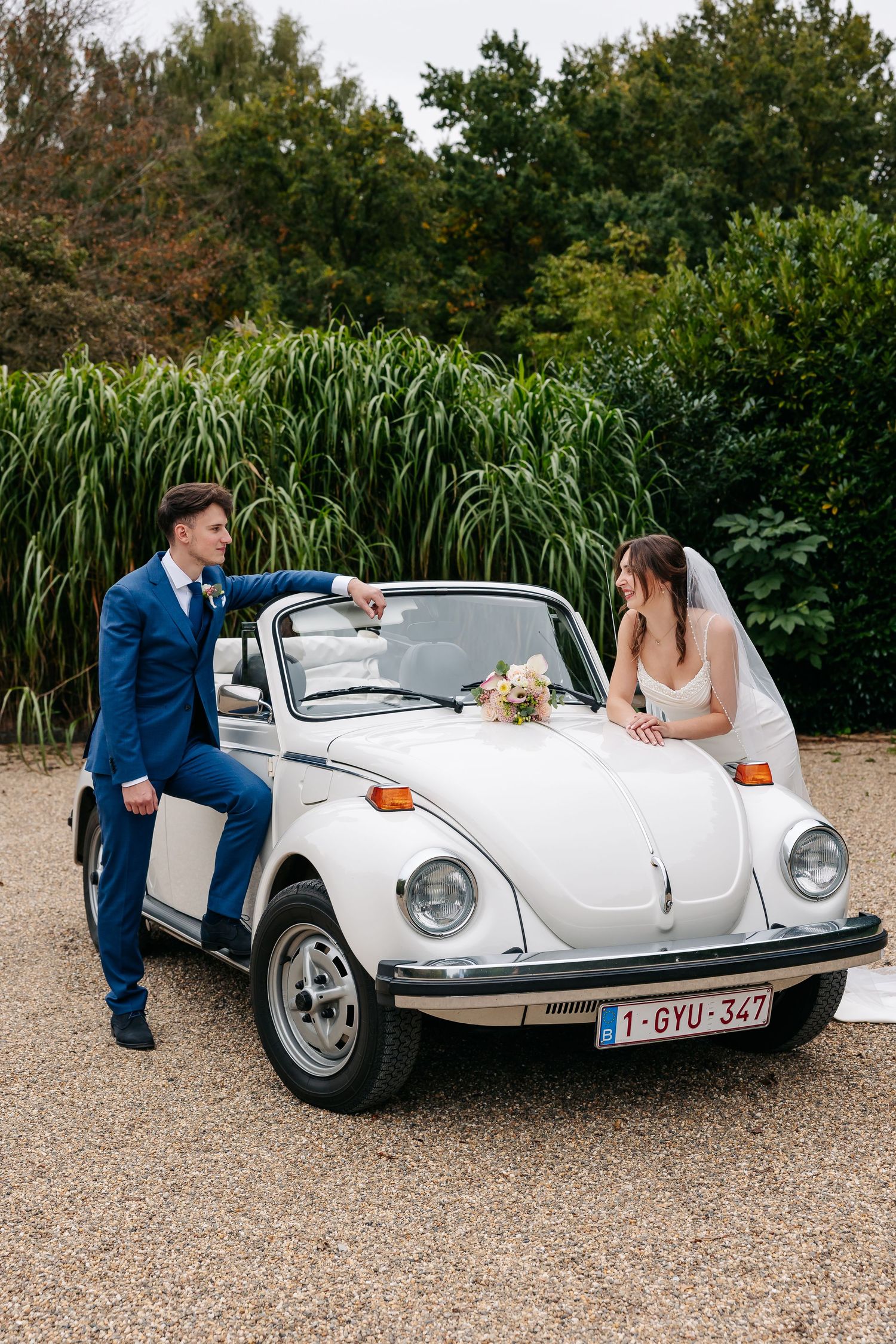 Bride and groom posing beside a classic white VW Beetle convertible with a bridal bouquet on hood.