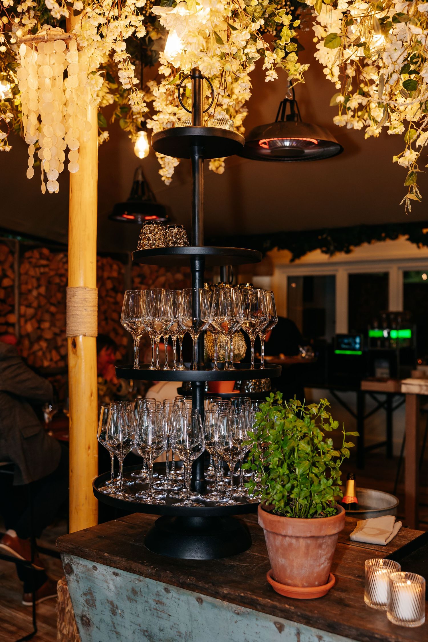Multi-tiered display of empty wine glasses on a rustic bar counter under warm fairy lights at a wedding venue.