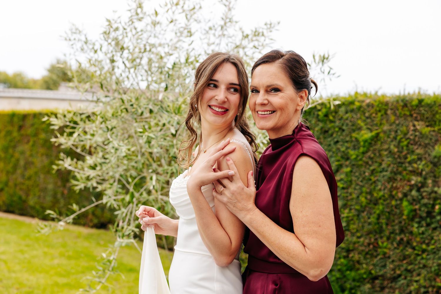 Smiling bride in white gown poses with a woman in a burgundy dress beside an olive tree.