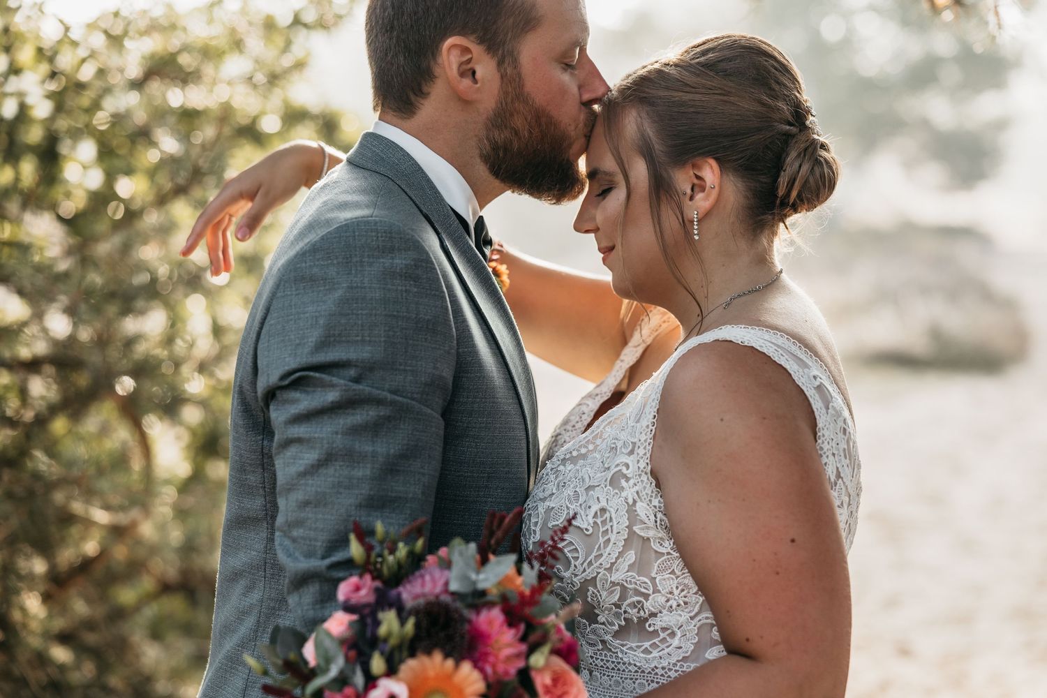 Groom gently kisses bride's forehead while she holds a bright floral bouquet in outdoor wedding photo.