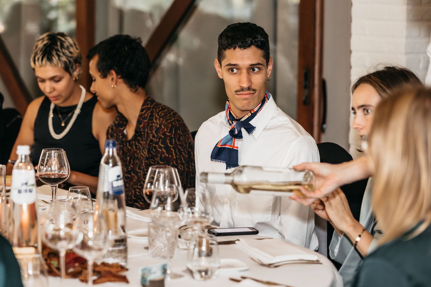 Guests seated at an elegant dinner table being served wine by a waiter in a bow tie.