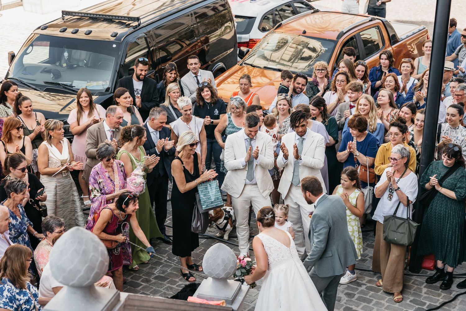Overhead view of joyful wedding guests celebrating with bride and groom outside venue near parked black van.