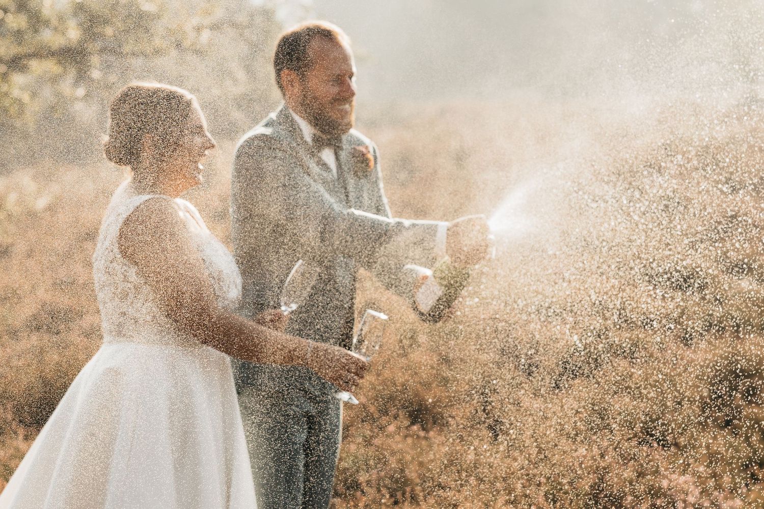 Joyful couple drenched in champagne spray in golden heathland during outdoor wedding celebration.
