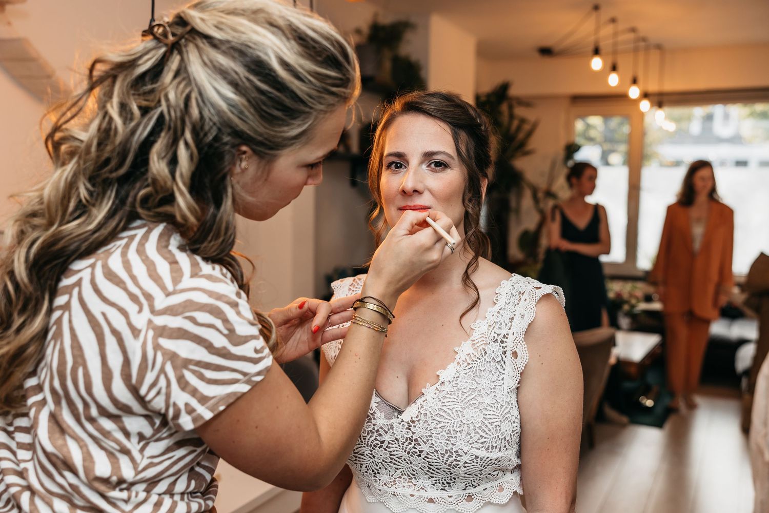 Makeup artist applying finishing touches to a bride in a white lace wedding dress before her wedding day.