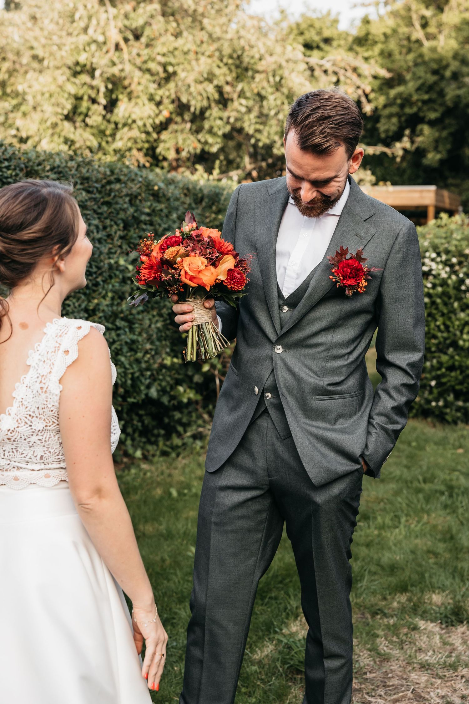 Groom in gray three-piece suit admiring red and orange bridal bouquet in outdoor garden.