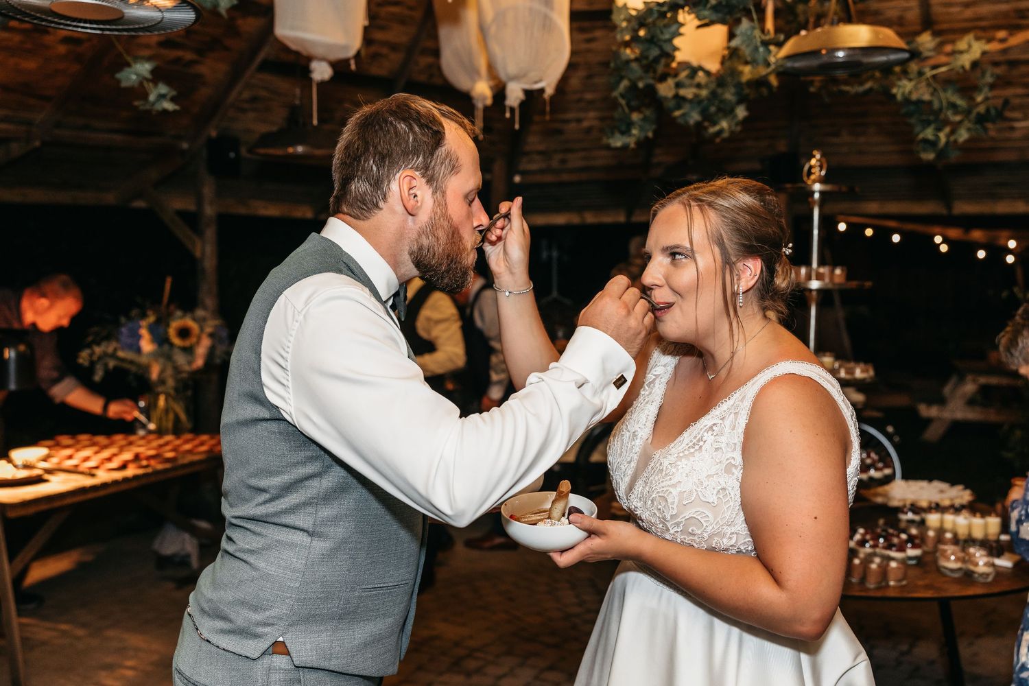 Groom feeding bride a sweet treat at rustic indoor wedding reception with bokeh lights.