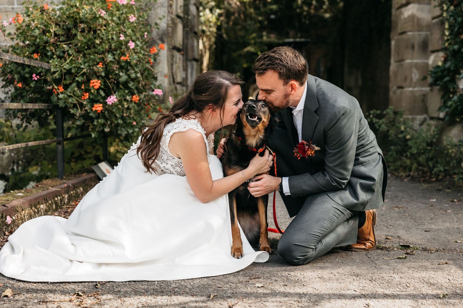 Bride and groom sitting on ground sharing a tender moment with their dog in a garden setting.