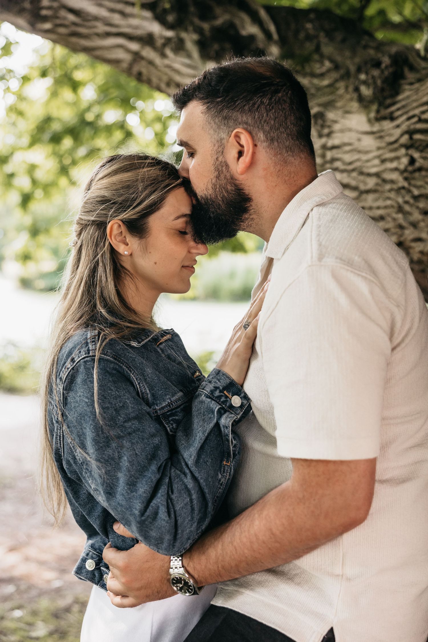 Romantic couple sharing an intimate moment under a tree, woman in denim jacket embracing bearded man.