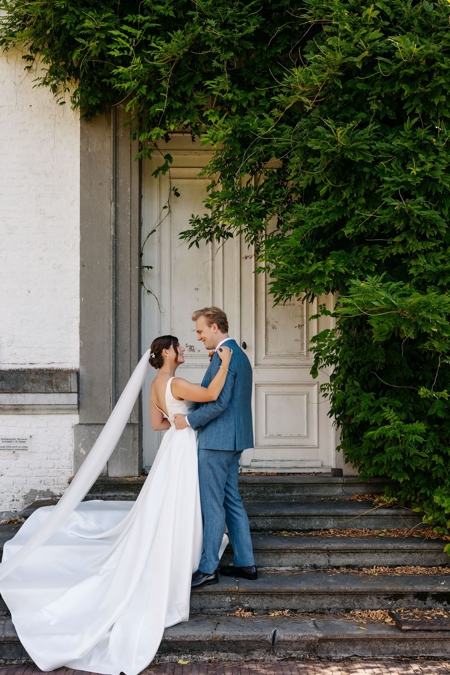 Bride and groom embracing at ornate white door surrounded by lush green ivy during wedding portraits.