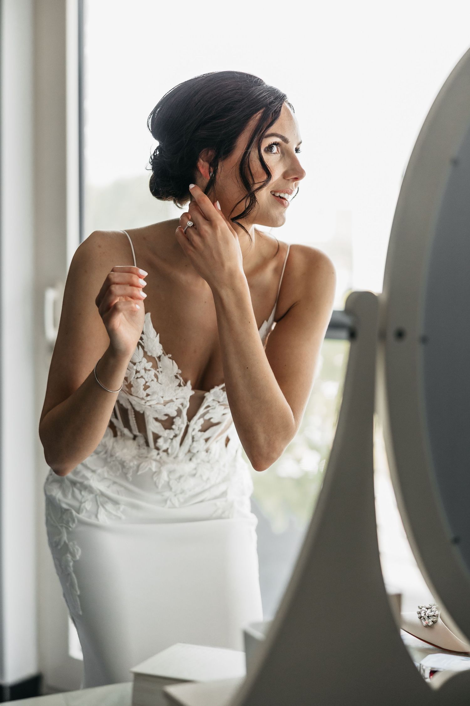 Radiant bride in lace wedding dress putting on earrings near a mirror.