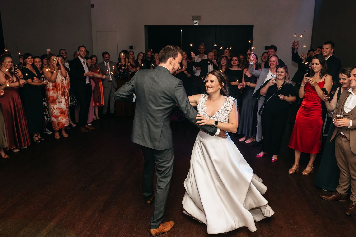 Bride and groom share first dance surrounded by guests holding sparklers in a modern reception venue.