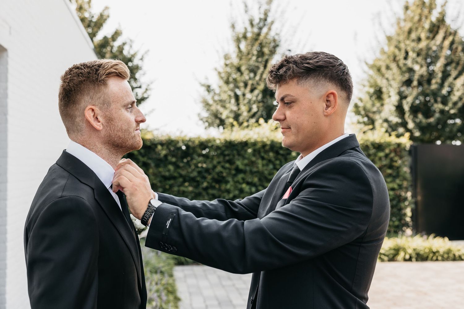 A groomsman carefully adjusts the tie of another man in a black suit outdoors.