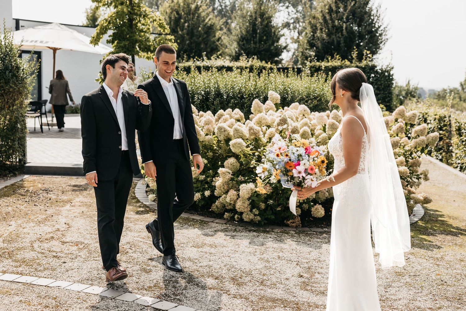 Two groomsmen in black suits approaching bride holding colorful bouquet near blooming hydrangeas at wedding.