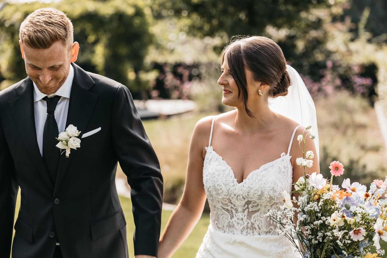 Bride and groom walking hand in hand through a sunlit garden, bride holding a colorful wildflower bouquet.