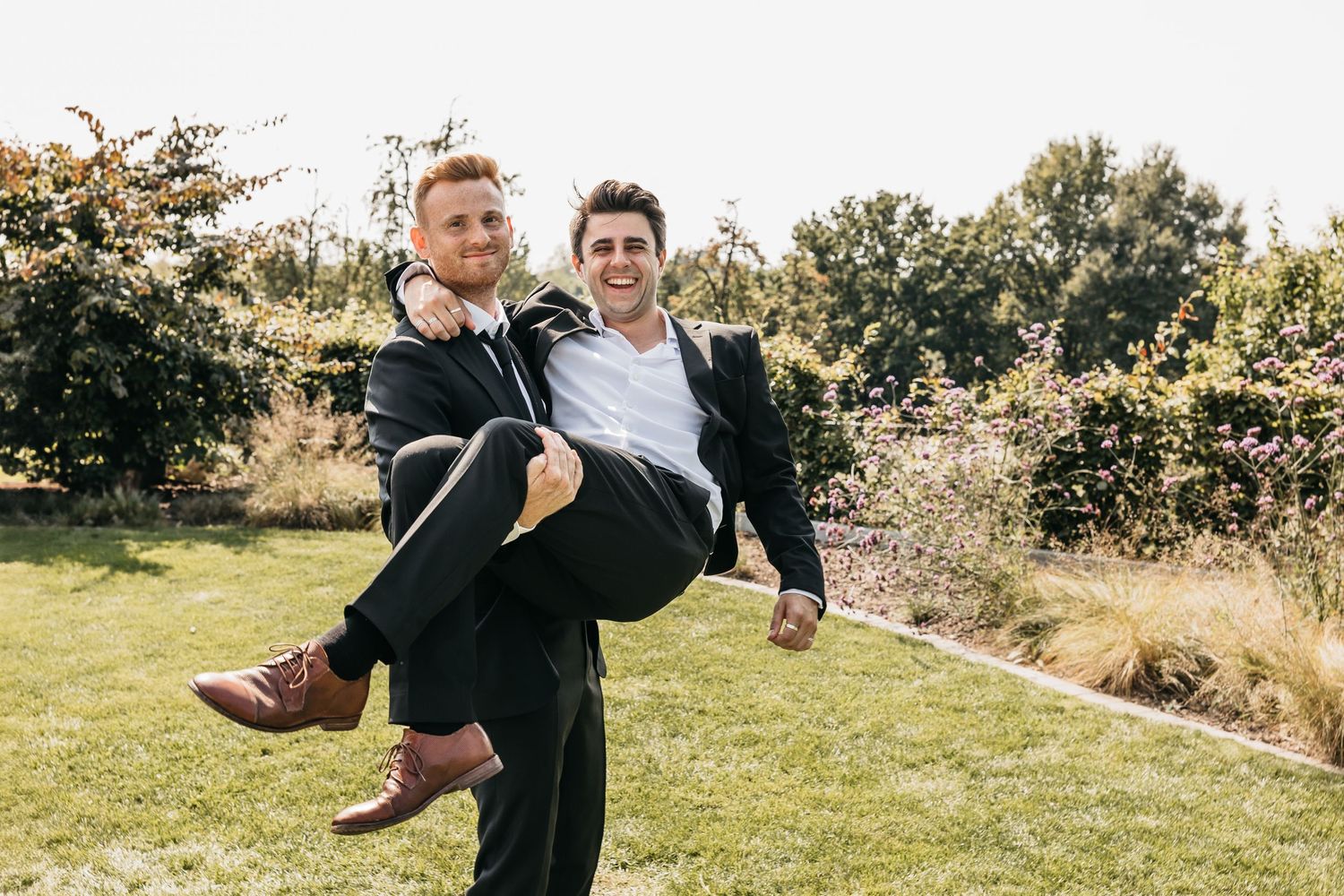 Groomsman carrying groom in arms while laughing together in garden at wedding.
