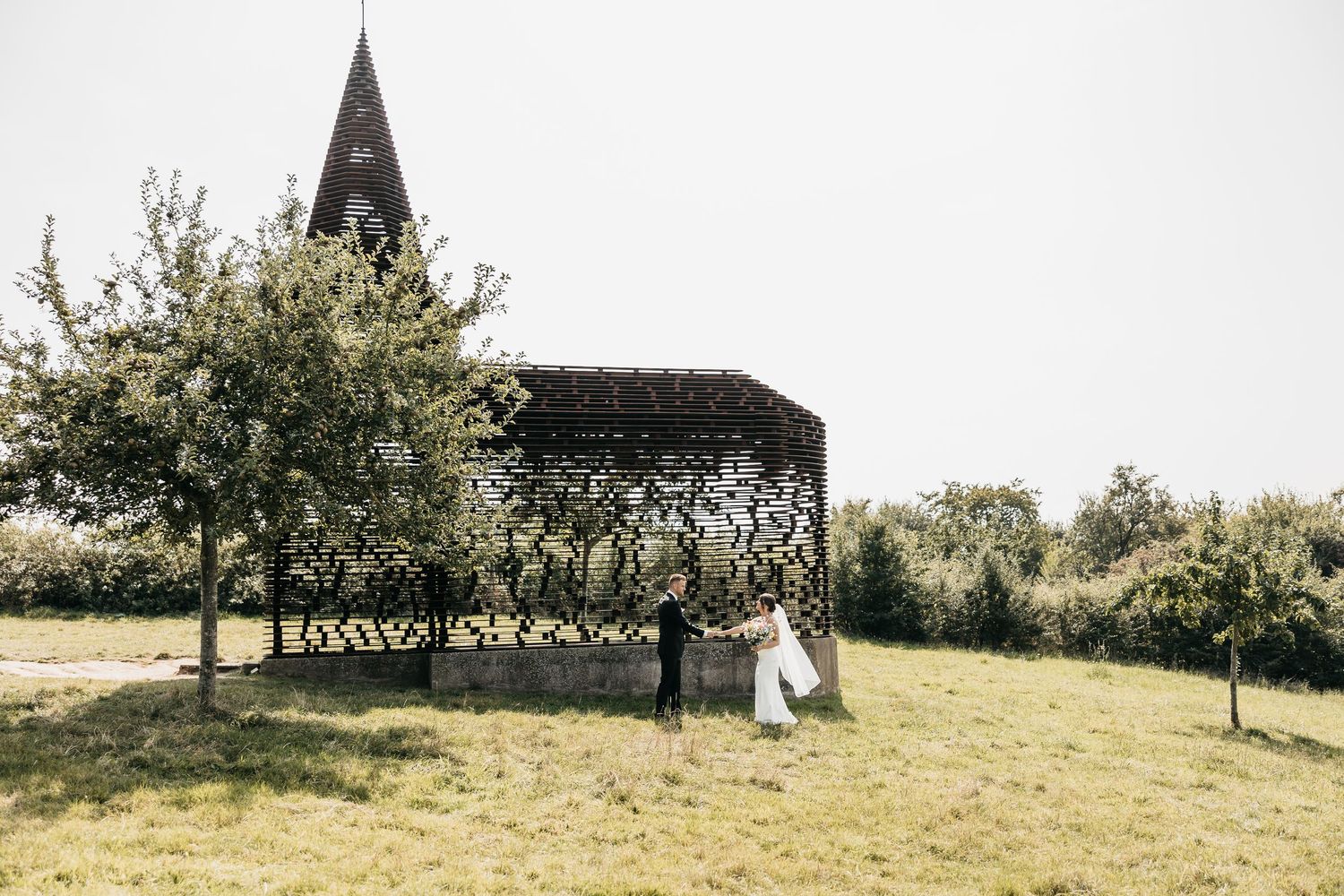 Bride and groom stand together near a unique wooden chapel structure surrounded by trees in an open field.