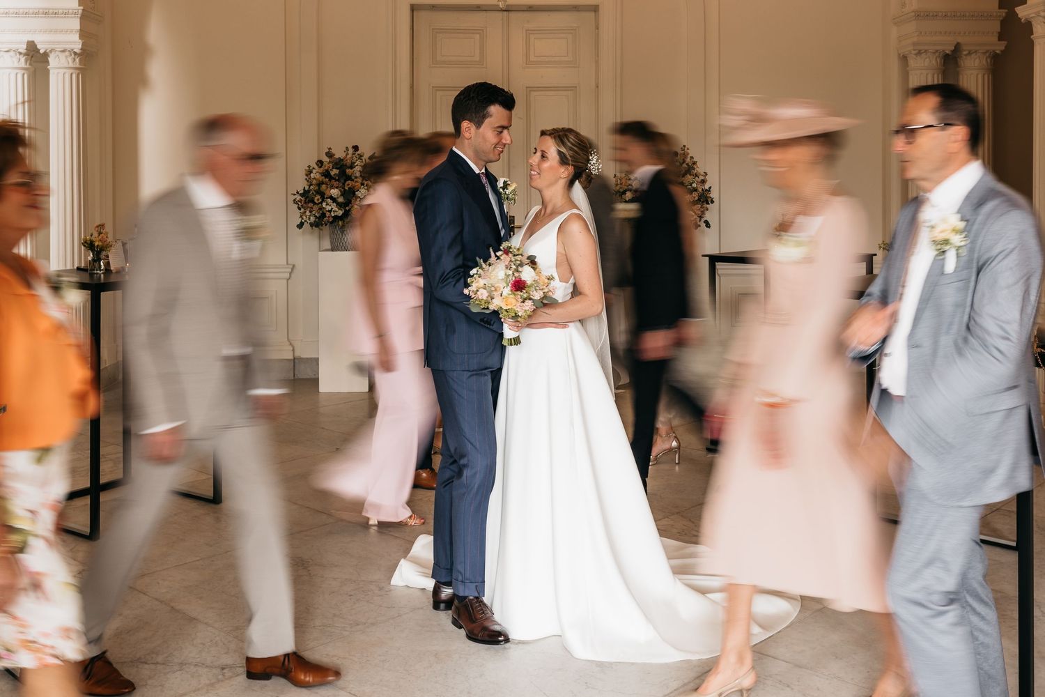 Bride and groom share a romantic moment as wedding guests blur past them in an elegant white venue.