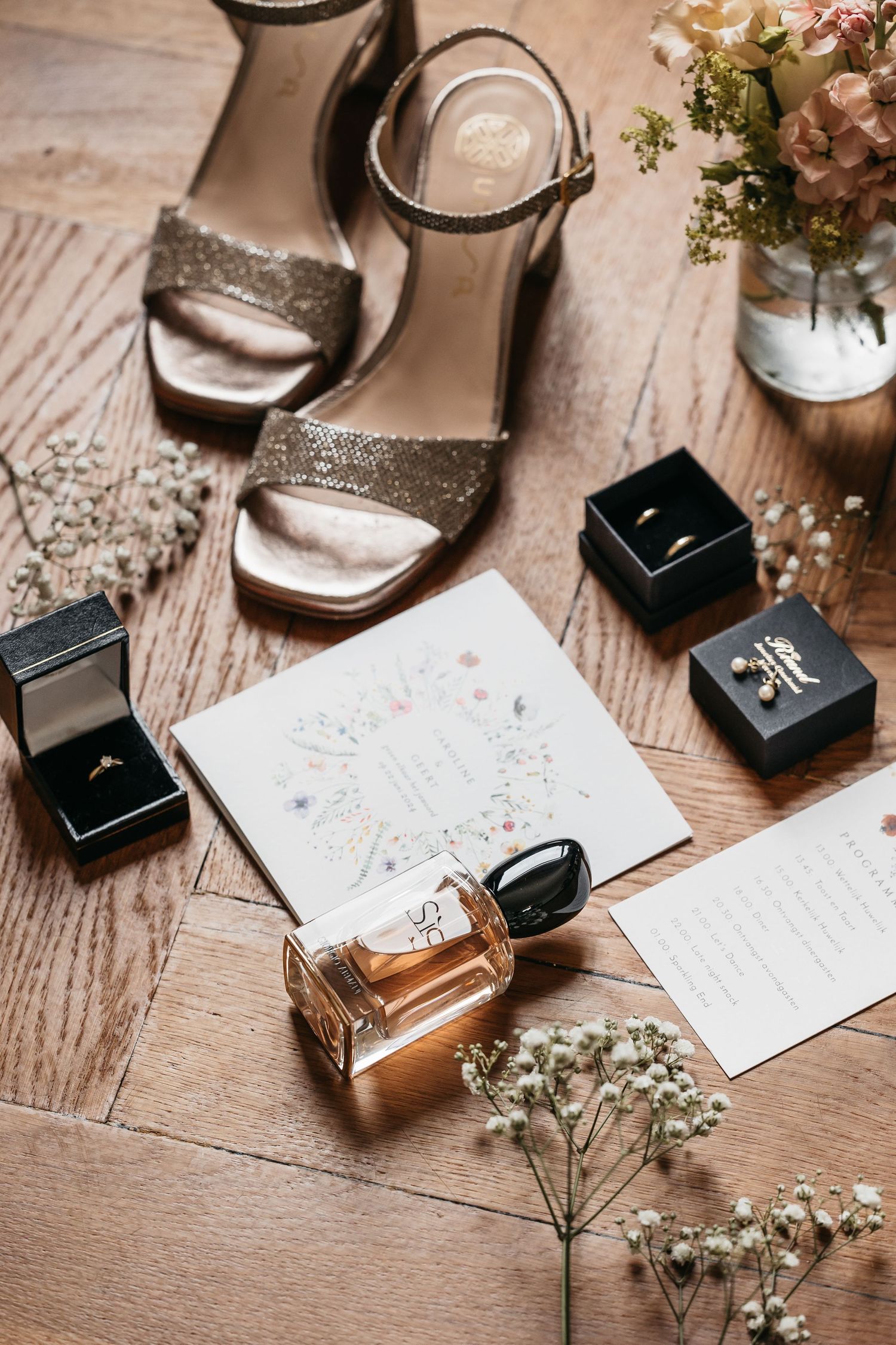 Wedding flat lay featuring glitter heels, rings, perfume, floral invitation, and dried flowers on wood.