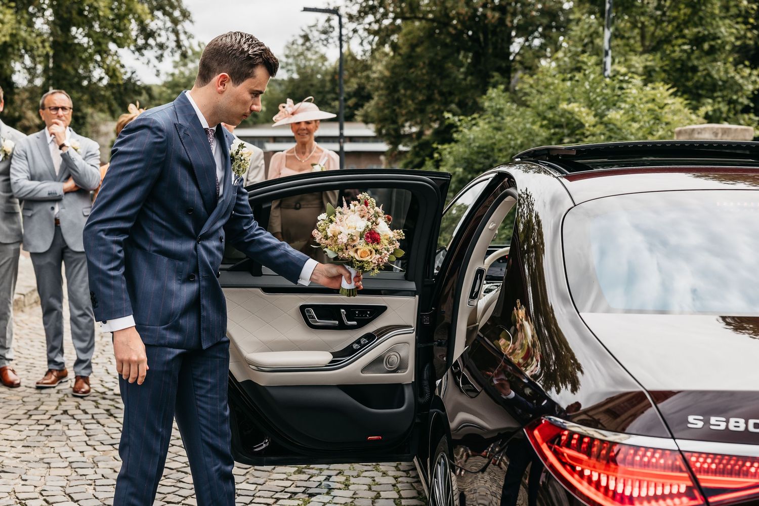 Groom in navy suit opening car door for bride holding bouquet outside a church on cobblestone street.