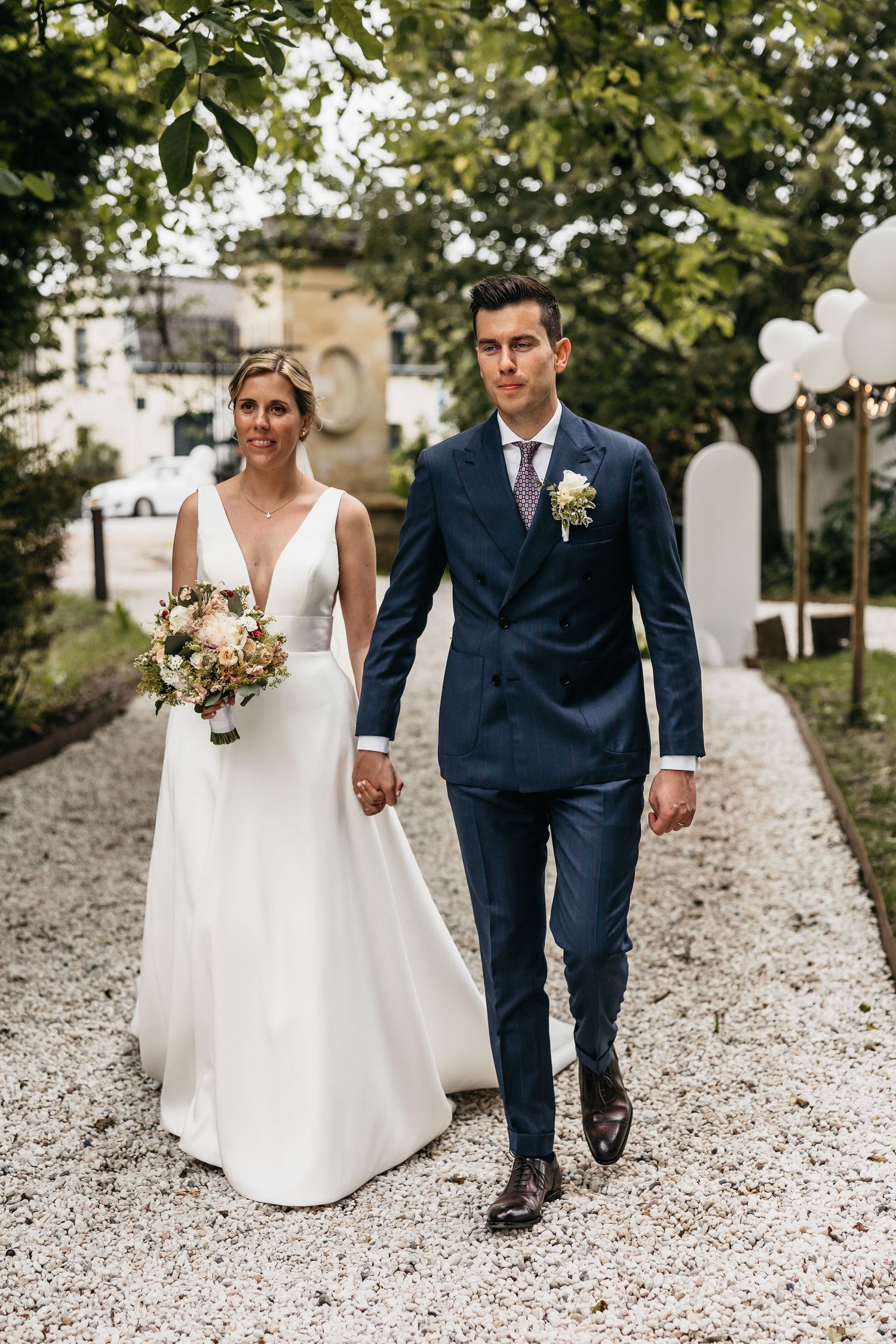 Elegant bride and groom walking hand in hand along gravel path with white balloon decorations.