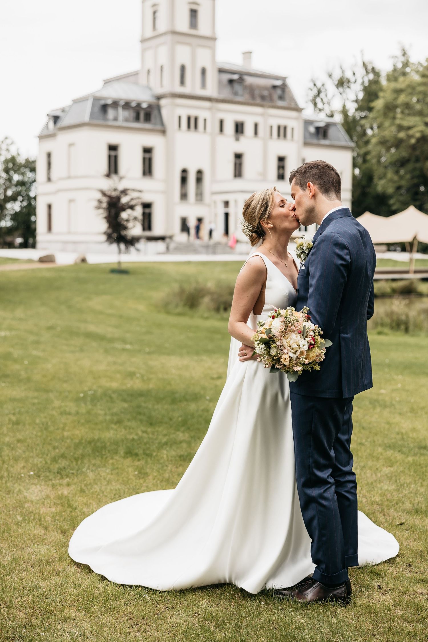Bride and groom kissing in front of a grand white chateau on a lush green lawn.