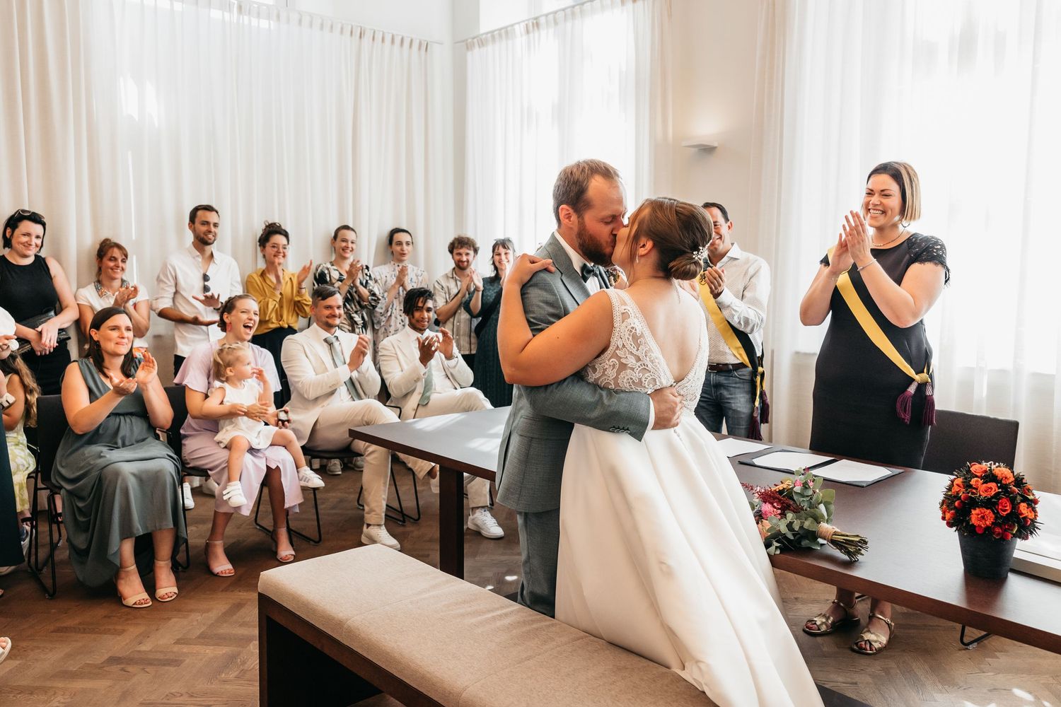 Newlyweds share romantic first kiss as guests cheer and clap at wedding ceremony.