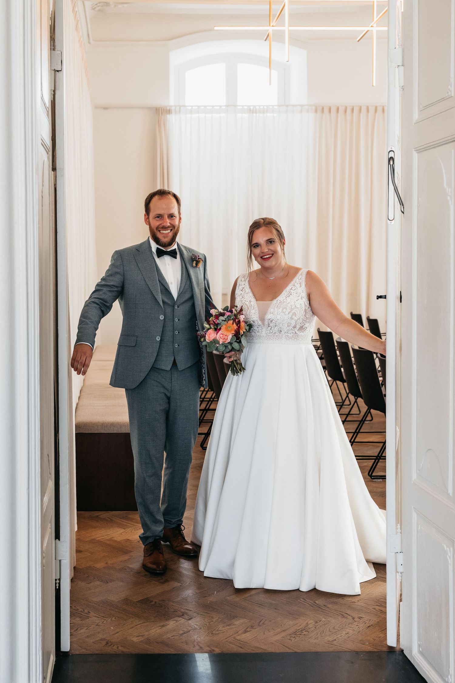 Happy newlyweds walking through white double doors into wedding ceremony room with chairs and herringbone floor.