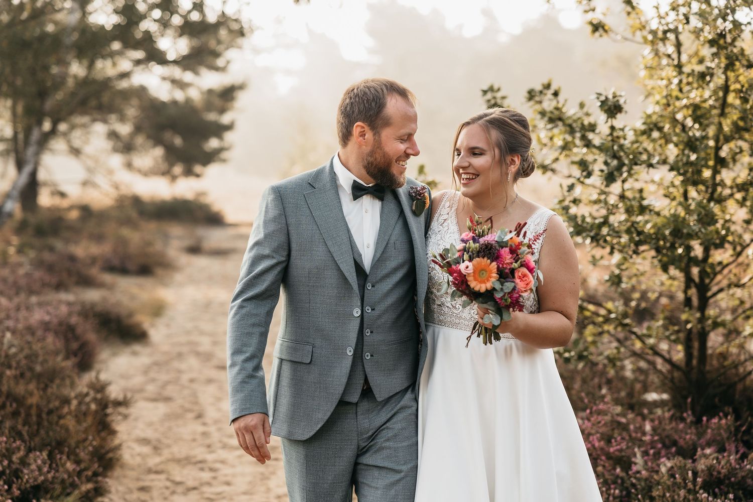 Happy bride and groom strolling through a misty heathland landscape, bride holding a colorful bouquet.