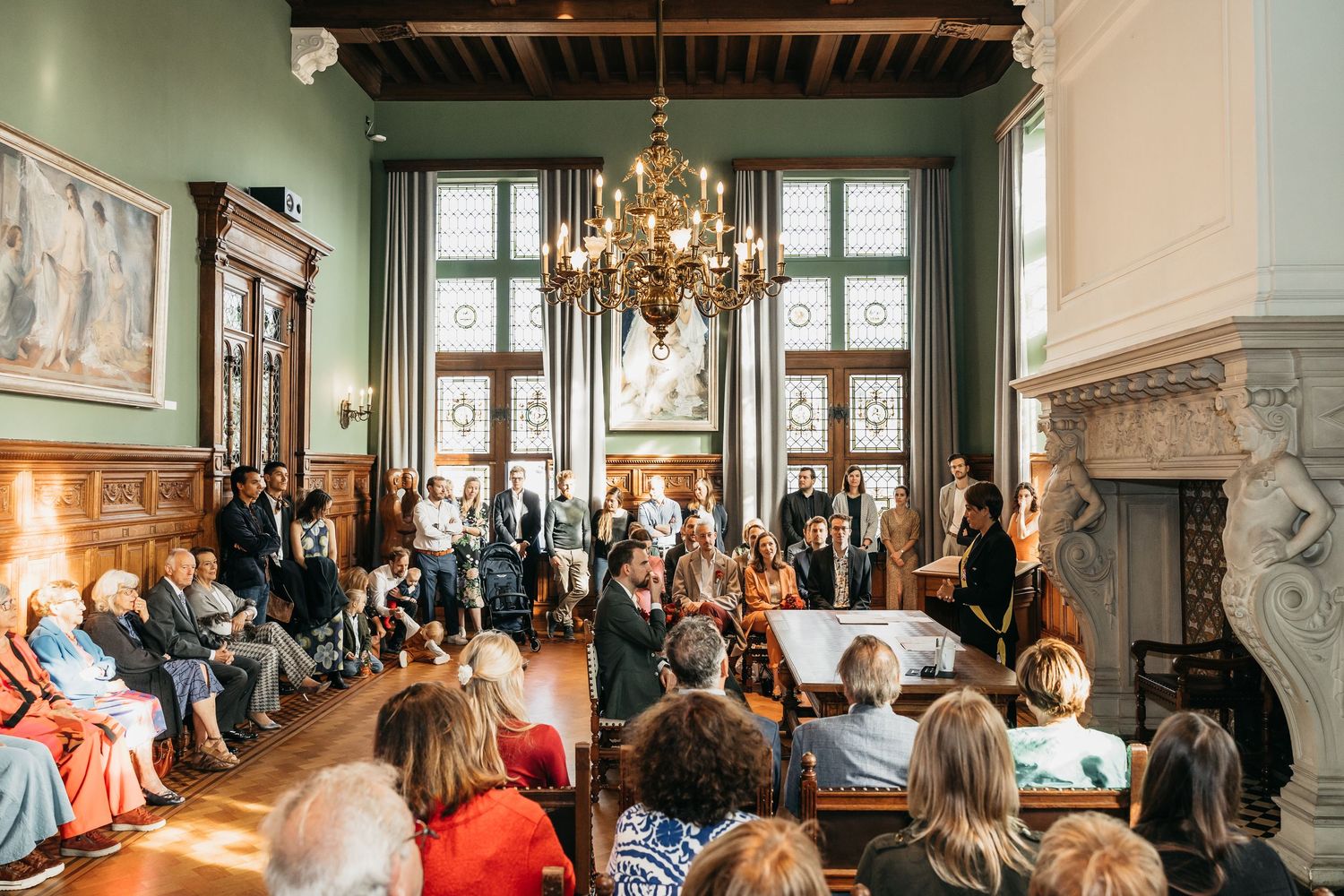 Sunlit wedding ceremony in a classic European hall with chandelier, guests gathered around the couple.